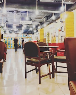 An indoor space with several wooden chairs and tables arranged in rows. The flooring is shiny and reflective, and the ceiling features many exposed pipes and lights. There are two people walking towards the far end of the room where a set of large windows and glass doors is located. The walls are adorned with decorations and there are some potted plants.