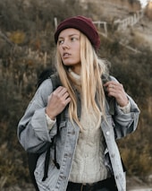 A person is standing outdoors, wearing a maroon beanie, a light-colored sweater, and a denim jacket. They have long blonde hair and are holding the straps of a backpack. The background is composed of an out-of-focus natural setting with earthy tones.