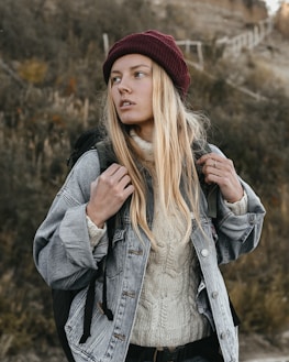 A person is standing outdoors, wearing a maroon beanie, a light-colored sweater, and a denim jacket. They have long blonde hair and are holding the straps of a backpack. The background is composed of an out-of-focus natural setting with earthy tones.