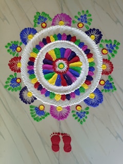 An intricate and vibrant rangoli design featuring concentric circles filled with colorful patterns, surrounded by floral motifs made of different colors including green, blue, yellow, red, and purple. The design is crafted on a light-colored, possibly marble floor, and includes a pair of red footprints at the bottom, suggesting a traditional or cultural significance.