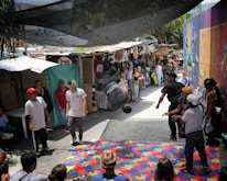 A group of people gather in an outdoor market area with various stalls and colorful graffiti on the walls. In the center, a person is performing a backflip on a mat made of puzzle pieces. Several people watch the performer, some wearing casual clothing and hats. The scene is lively and dynamic, with a mix of bright colors in the surroundings.