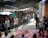 A group of people gather in an outdoor market area with various stalls and colorful graffiti on the walls. In the center, a person is performing a backflip on a mat made of puzzle pieces. Several people watch the performer, some wearing casual clothing and hats. The scene is lively and dynamic, with a mix of bright colors in the surroundings.