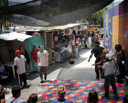 A group of people gather in an outdoor market area with various stalls and colorful graffiti on the walls. In the center, a person is performing a backflip on a mat made of puzzle pieces. Several people watch the performer, some wearing casual clothing and hats. The scene is lively and dynamic, with a mix of bright colors in the surroundings.