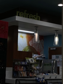 A snack bar area inside a store featuring a variety of packaged chips and snacks neatly arranged on a shelf. Above the shelf, there is a large, lit-up graphic with an artistic design, and two modern pendant lights hang from the ceiling. The word 'refresh' is prominently displayed at the top. The background includes a blue wall and a door with some posters or notices on it.