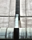 The image depicts an industrial structure featuring a large metal duct or vent running vertically against a concrete wall. The surface of the duct has a worn, metallic appearance with visible weathering and age. The concrete wall is segmented by horizontal and vertical lines, giving a geometric and symmetrical look.
