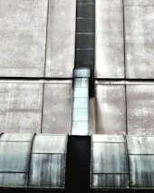 The image depicts an industrial structure featuring a large metal duct or vent running vertically against a concrete wall. The surface of the duct has a worn, metallic appearance with visible weathering and age. The concrete wall is segmented by horizontal and vertical lines, giving a geometric and symmetrical look.