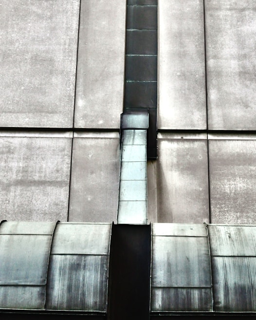 The image depicts an industrial structure featuring a large metal duct or vent running vertically against a concrete wall. The surface of the duct has a worn, metallic appearance with visible weathering and age. The concrete wall is segmented by horizontal and vertical lines, giving a geometric and symmetrical look.