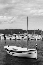 A black and white photograph featuring a small sailboat anchored on calm waters with a backdrop of numerous boats docked at a marina. The scene is set against a mountainous landscape under a cloudy sky.