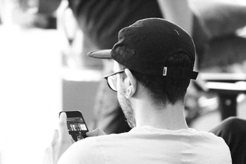 A person wearing a cap and glasses is viewed from behind, holding a smartphone in one hand. The setting appears to be informal, possibly indoors, with blurred elements in the background suggesting movement or other people.