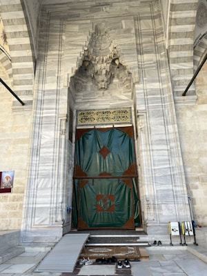 A grand, ornately decorated stone entrance with intricate marble carvings and patterns leads to a large door covered with a green and brown protective covering. Arabic script is visible above the door. Shoes are placed on a mat in front of the entrance, alongside a ramp for accessibility.