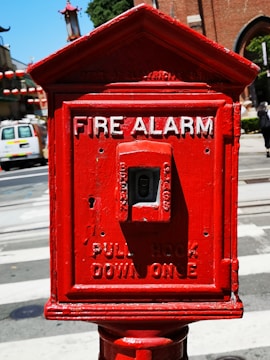 A vintage red fire alarm box with white lettering stands prominently on a street corner. The words 'Fire Alarm' are clearly visible at the top, with instructions to 'Pull Hook Down Once' below. Background elements include a road, a parked van, and buildings with architectural details such as awnings and lanterns.