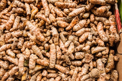 A large pile of fresh turmeric roots is displayed in a chaotic arrangement. The roots have a rough, textured surface with shades of brown and orange, indicating their raw, unprocessed state. Some of the roots are broken or split, revealing their vibrant orange interior.