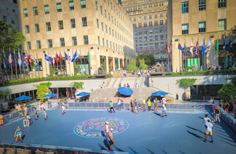 A vibrant outdoor scene featuring a roller skating rink surrounded by buildings with numerous national flags displayed. People are skating on the rink, while others watch or relax under blue umbrellas. The setting is a busy urban plaza with a central staircase leading up to a higher level, where more people are gathered.