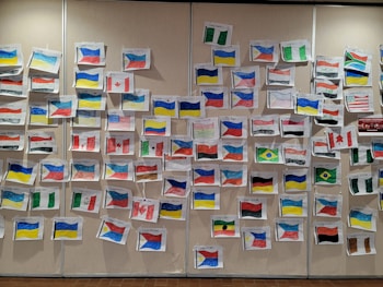 A wall is covered with handmade drawings of various national flags, each labeled with a country name. The drawings are arranged in a scattered pattern and vary in size. The flags include multiple colors, with prominent ones like red, blue, yellow, and green. This display likely represents a multicultural event or educational project highlighting diversity.