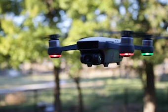 A drone is hovering mid-air with trees and blurred greenery in the background. The drone is equipped with bright red and green lights on its arms and appears to have a camera on its front. The image is captured during daylight, providing a clear view of the drone against the natural backdrop.