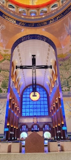 An interior of a grand cathedral with a large, prominent cross suspended in front of a vibrant blue stained glass window. The ceiling is adorned with intricate mosaics and painted murals, featuring religious and natural motifs. Soft lighting adds to the serene ambiance, with symmetrical arches leading to the altar.