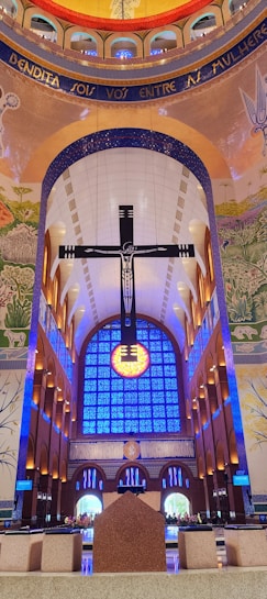 An interior of a grand cathedral with a large, prominent cross suspended in front of a vibrant blue stained glass window. The ceiling is adorned with intricate mosaics and painted murals, featuring religious and natural motifs. Soft lighting adds to the serene ambiance, with symmetrical arches leading to the altar.
