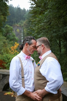 Two men in matching attire, consisting of white shirts with pink bow ties and suspenders, stand close together in a serene outdoor setting, surrounded by lush green trees. They are holding hands and touching foreheads, exuding a sense of intimacy and love. A waterfall is visible in the blurred background, enhancing the peaceful ambiance.
