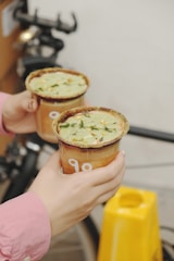 Two hands holding cups filled with a green beverage topped with herbs. The cups are brown with sugar-rimmed edges. A bicycle and a yellow object are in the background.