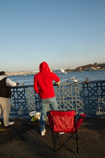 A person in a red hoodie is standing by a waterfront, holding a fishing rod. Another person is fishing beside them. A red folding chair is placed nearby on the pavement. A large body of water with a few boats and a distant cityscape can be seen in the background.
