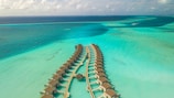 An aerial view of overwater bungalows arranged in a curved pattern over a turquoise sea with variations in water color indicating shallow waters and coral reefs. The bungalows are linked by a wooden walkway, and there are scattered clouds in the sky.