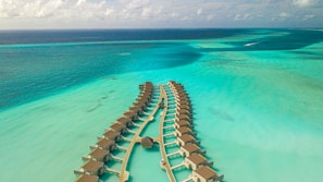 An aerial view of overwater bungalows arranged in a curved pattern over a turquoise sea with variations in water color indicating shallow waters and coral reefs. The bungalows are linked by a wooden walkway, and there are scattered clouds in the sky.