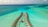 An aerial view of overwater bungalows arranged in a curved pattern over a turquoise sea with variations in water color indicating shallow waters and coral reefs. The bungalows are linked by a wooden walkway, and there are scattered clouds in the sky.