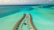 An aerial view of overwater bungalows arranged in a curved pattern over a turquoise sea with variations in water color indicating shallow waters and coral reefs. The bungalows are linked by a wooden walkway, and there are scattered clouds in the sky.