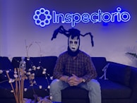 A person wearing a spooky mask sits on a dark sofa under a neon blue sign that reads 'Inspectorio'. The mask features black and white makeup with spider-like appendages on the head. There are decorative items, including a small skeleton figure and dried plants, on the table in front and another smaller skeleton on the sofa.
