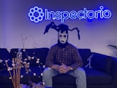 A person wearing a spooky mask sits on a dark sofa under a neon blue sign that reads 'Inspectorio'. The mask features black and white makeup with spider-like appendages on the head. There are decorative items, including a small skeleton figure and dried plants, on the table in front and another smaller skeleton on the sofa.