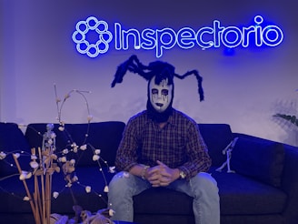 A person wearing a spooky mask sits on a dark sofa under a neon blue sign that reads 'Inspectorio'. The mask features black and white makeup with spider-like appendages on the head. There are decorative items, including a small skeleton figure and dried plants, on the table in front and another smaller skeleton on the sofa.