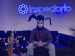 A person wearing a spooky mask sits on a dark sofa under a neon blue sign that reads 'Inspectorio'. The mask features black and white makeup with spider-like appendages on the head. There are decorative items, including a small skeleton figure and dried plants, on the table in front and another smaller skeleton on the sofa.