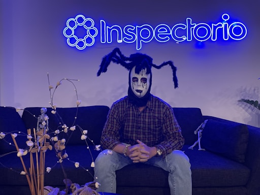 A person wearing a spooky mask sits on a dark sofa under a neon blue sign that reads 'Inspectorio'. The mask features black and white makeup with spider-like appendages on the head. There are decorative items, including a small skeleton figure and dried plants, on the table in front and another smaller skeleton on the sofa.