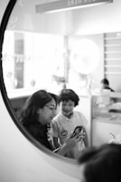 A woman and a child are looking at a smartphone together, captured through a circular frame. The setting seems casual and light-hearted, possibly in a cafe or an informal environment. The woman is wearing glasses and has dark hair, while the child is smiling and wearing a light-colored shirt. In the background, there are blurred elements like a counter and a bright light fixture, suggesting an indoor space.