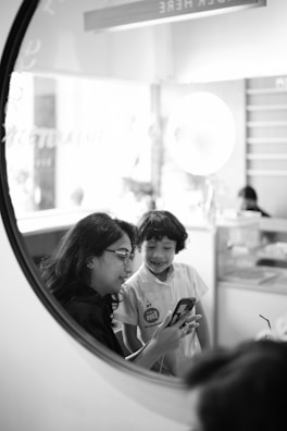 A woman and a child are looking at a smartphone together, captured through a circular frame. The setting seems casual and light-hearted, possibly in a cafe or an informal environment. The woman is wearing glasses and has dark hair, while the child is smiling and wearing a light-colored shirt. In the background, there are blurred elements like a counter and a bright light fixture, suggesting an indoor space.