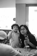 A black and white image featuring a woman and a child indoors. The child is wearing a school uniform and appears to be interacting with the woman who is holding a smartphone. Both are looking toward the camera with light coming from the window behind them.