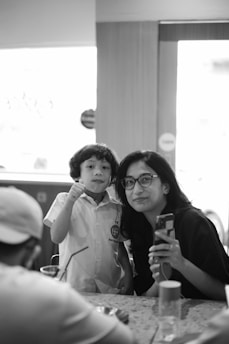 A black and white image featuring a woman and a child indoors. The child is wearing a school uniform and appears to be interacting with the woman who is holding a smartphone. Both are looking toward the camera with light coming from the window behind them.