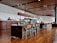 The interior of a modern cafe with a long wooden communal table and matching chairs in the center. The cafe has wooden flooring, a high ceiling with recessed lighting, and a wooden counter to the left with a staff member behind it. There are various items on the counter, including a menu board and some food displays. In the background, there is a refrigerator and some potted plants. The overall decor is minimalist and contemporary.