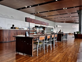 The interior of a modern cafe with a long wooden communal table and matching chairs in the center. The cafe has wooden flooring, a high ceiling with recessed lighting, and a wooden counter to the left with a staff member behind it. There are various items on the counter, including a menu board and some food displays. In the background, there is a refrigerator and some potted plants. The overall decor is minimalist and contemporary.