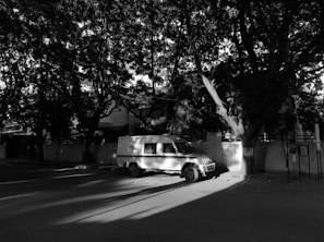 A black and white image features a parked security vehicle on a quiet street under the shade of large trees. The contrast between the sunlight and shadows gives the scene a dramatic appearance. The vehicle is positioned beside a wall, and the surrounding area appears calm and undisturbed.