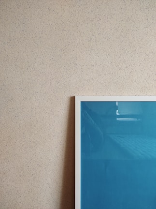 A textured beige wall is adorned with a framed artwork. The frame has a white border and contains a blue-colored piece. The photo captures a minimalist and modern aesthetic.