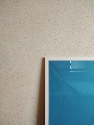 A textured beige wall is adorned with a framed artwork. The frame has a white border and contains a blue-colored piece. The photo captures a minimalist and modern aesthetic.