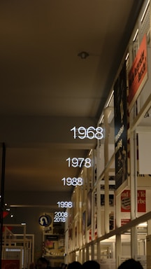 A corridor with a timeline display featuring illuminated years from 1968 to 2018. Posters and informational displays are mounted on the walls, indicating an exhibition or museum setting. The ceiling is dark, creating a contrast with the bright neon numbers.