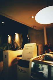 A cozy coffee shop setting with dim lighting featuring a coffee machine and a notice titled 'Travelator Blend'. The notice is placed on a jar labeled 'Arabica Beans', and there are leafy plants in the background. The warm ambiance is accentuated by soft lights and wooden surfaces.