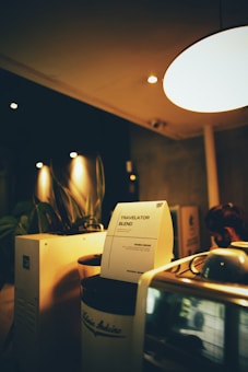 A cozy coffee shop setting with dim lighting featuring a coffee machine and a notice titled 'Travelator Blend'. The notice is placed on a jar labeled 'Arabica Beans', and there are leafy plants in the background. The warm ambiance is accentuated by soft lights and wooden surfaces.