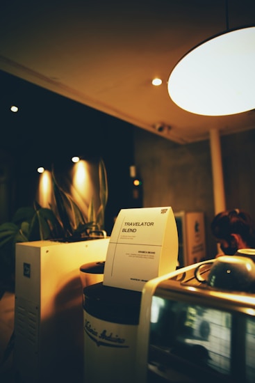 A cozy coffee shop setting with dim lighting featuring a coffee machine and a notice titled 'Travelator Blend'. The notice is placed on a jar labeled 'Arabica Beans', and there are leafy plants in the background. The warm ambiance is accentuated by soft lights and wooden surfaces.