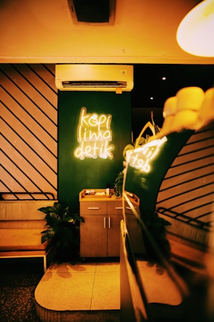 A cozy café interior with a neon sign displaying 'kopi lima detik' on a dark green wall. An air conditioner is mounted above the sign. There's a small wooden cabinet below the sign and a leather bench to the left. Potted plants add greenery, and warm ambient lighting creates a welcoming atmosphere.
