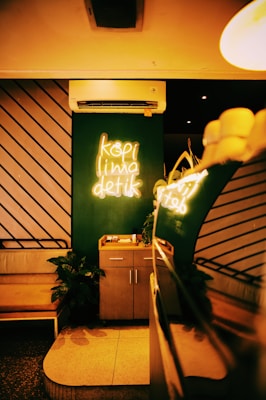 A cozy café interior with a neon sign displaying 'kopi lima detik' on a dark green wall. An air conditioner is mounted above the sign. There's a small wooden cabinet below the sign and a leather bench to the left. Potted plants add greenery, and warm ambient lighting creates a welcoming atmosphere.