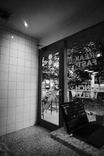 A monochrome photograph of a cafe interior with tiled walls and a door with glass panels. Outside the glass door, there is a person sitting on a stool, visible in the reflection. Inside the cafe, a laptop rests on a stone-textured table, next to a cup.