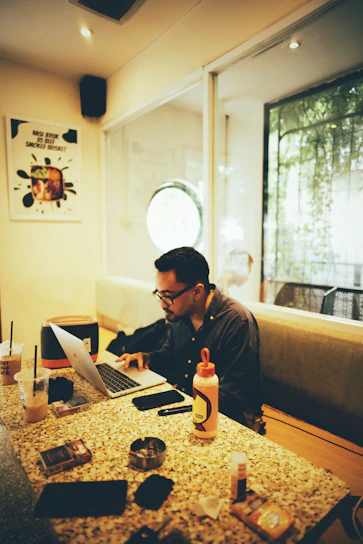 A candid photo of a smiling man sitting at a cozy café table with a laptop and coffee, sunlight streaming through the window.